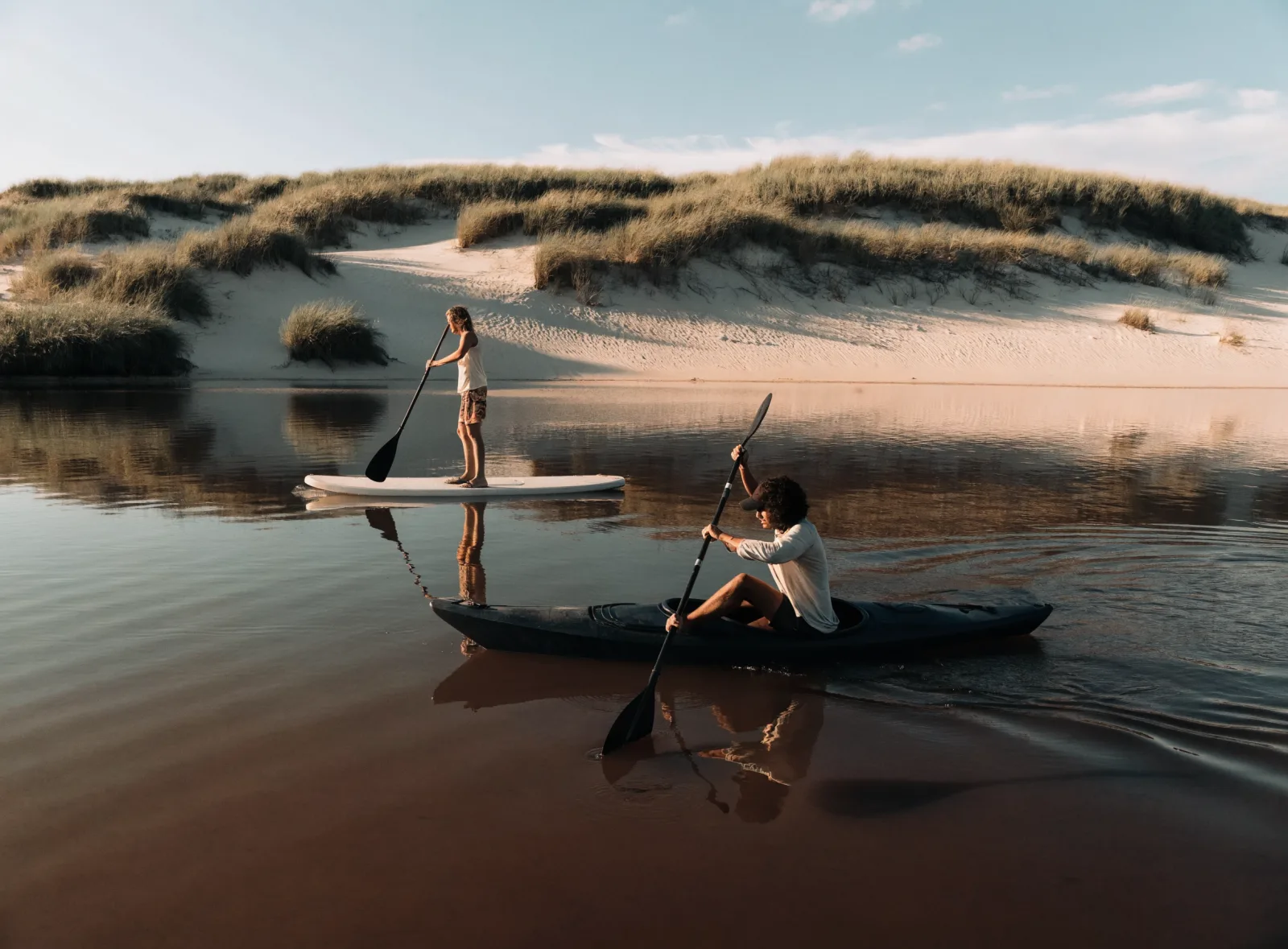 Paddlers crossing the dark water of a coastal dune lake toward high white dunes in late light