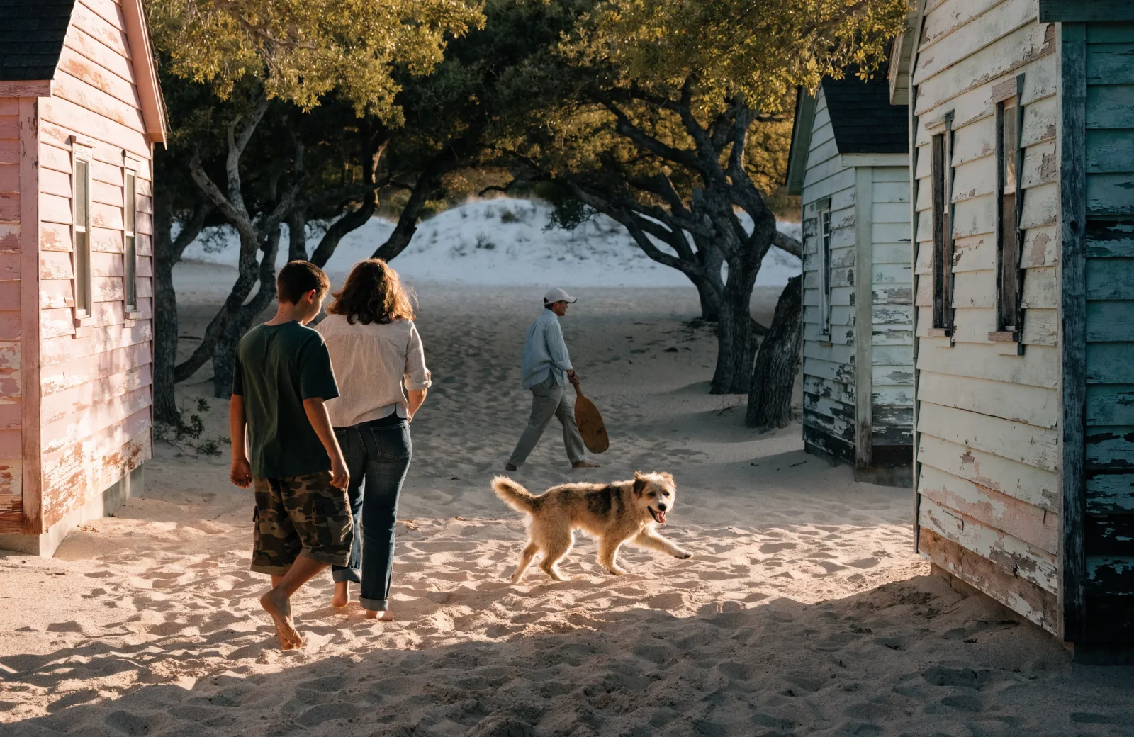 Family walking a sandy lane between weathered cottages and oaks toward the dunes, dog trotting ahead